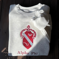 Load image into Gallery viewer, Light gray sweatshirt featuring a large, embroidered shield of the Alpha Phi sorority in a deep red and silver, prominently displayed on the front. The shield includes a diagonal band with silver stars.