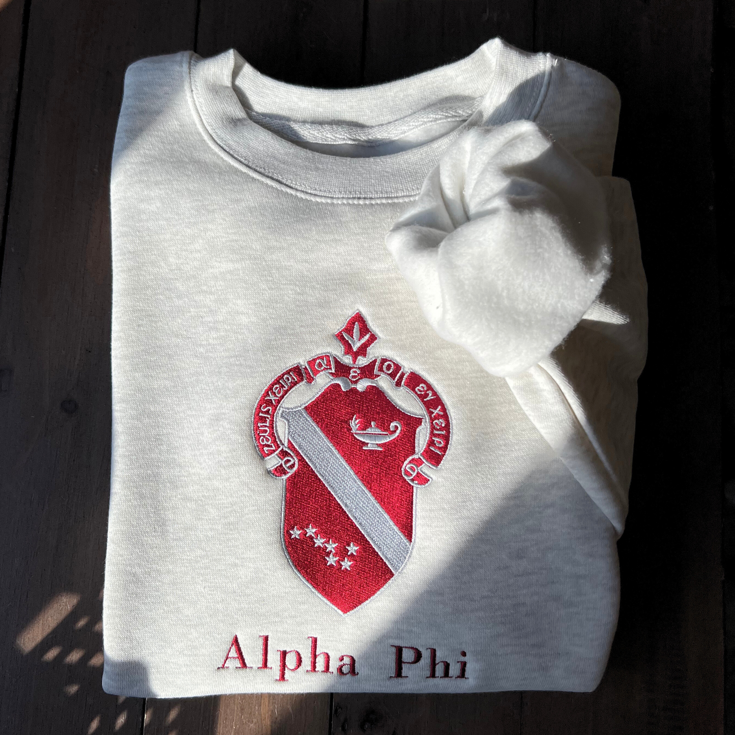 Light gray sweatshirt featuring a large, embroidered shield of the Alpha Phi sorority in a deep red and silver, prominently displayed on the front. The shield includes a diagonal band with silver stars.