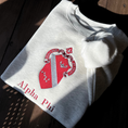 Load image into Gallery viewer, Light gray sweatshirt featuring a large, embroidered shield of the Alpha Phi sorority in a deep red and silver, prominently displayed on the front. The shield includes a diagonal band with silver stars.