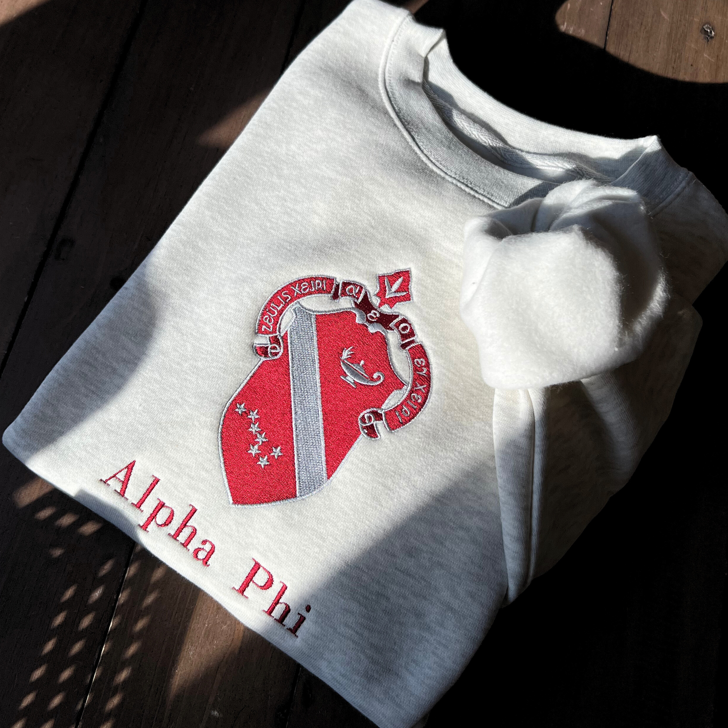 Light gray sweatshirt featuring a large, embroidered shield of the Alpha Phi sorority in a deep red and silver, prominently displayed on the front. The shield includes a diagonal band with silver stars.