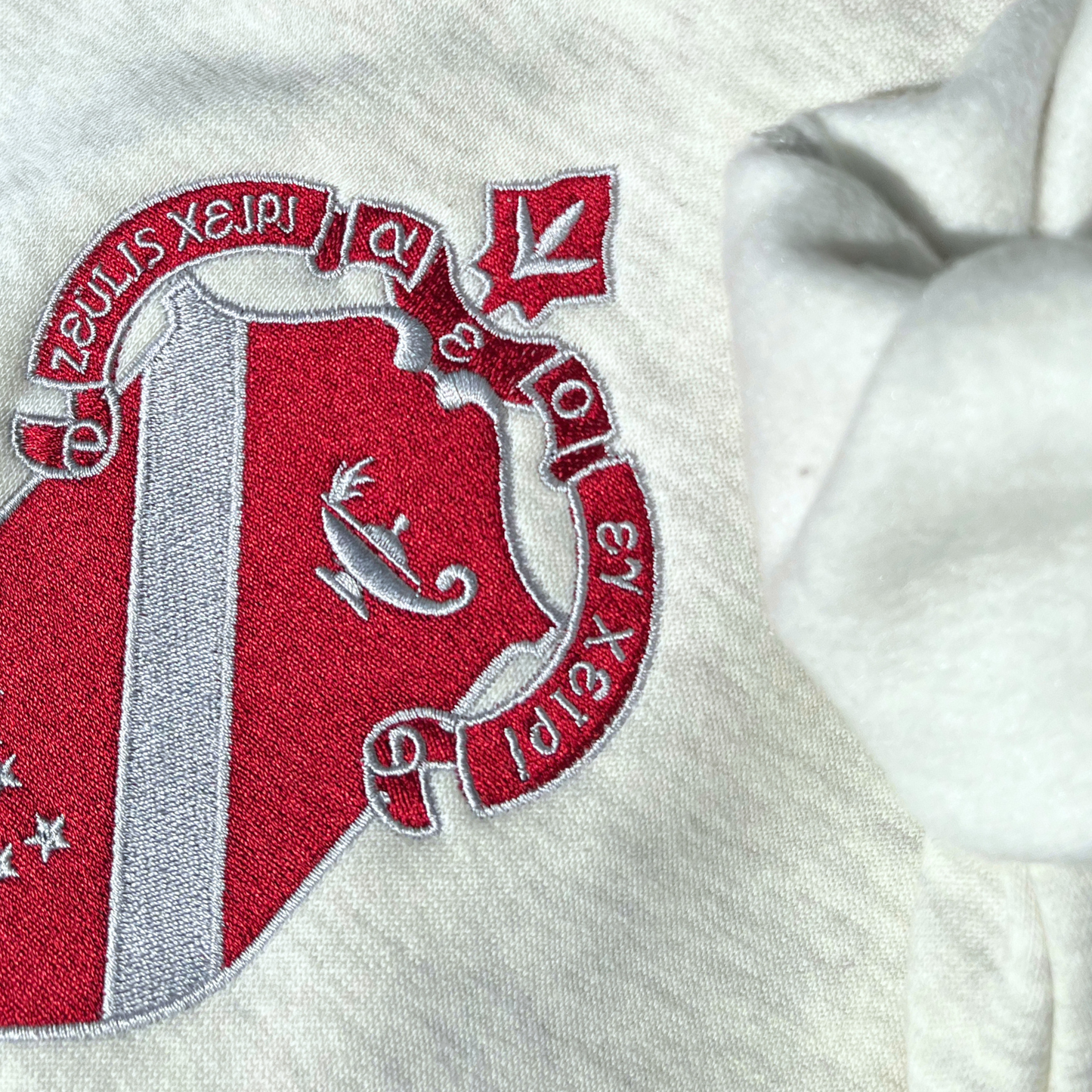 A detailed view of a light gray sweatshirt featuring a large, embroidered shield of the Alpha Phi sorority in a deep red and silver, prominently displayed on the front. The shield includes a diagonal band with silver stars.