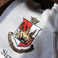 Load image into Gallery viewer, Detailed view of a light gray crewneck sweatshirt laid flat on a wooden surface, showcasing the Alpha Sigma Alpha sorority crest embroidered in the center. The crest displays a red shield with a golden phoenix, topped by a crown, and surrounded by a scroll bearing the sorority's Greek motto and name.