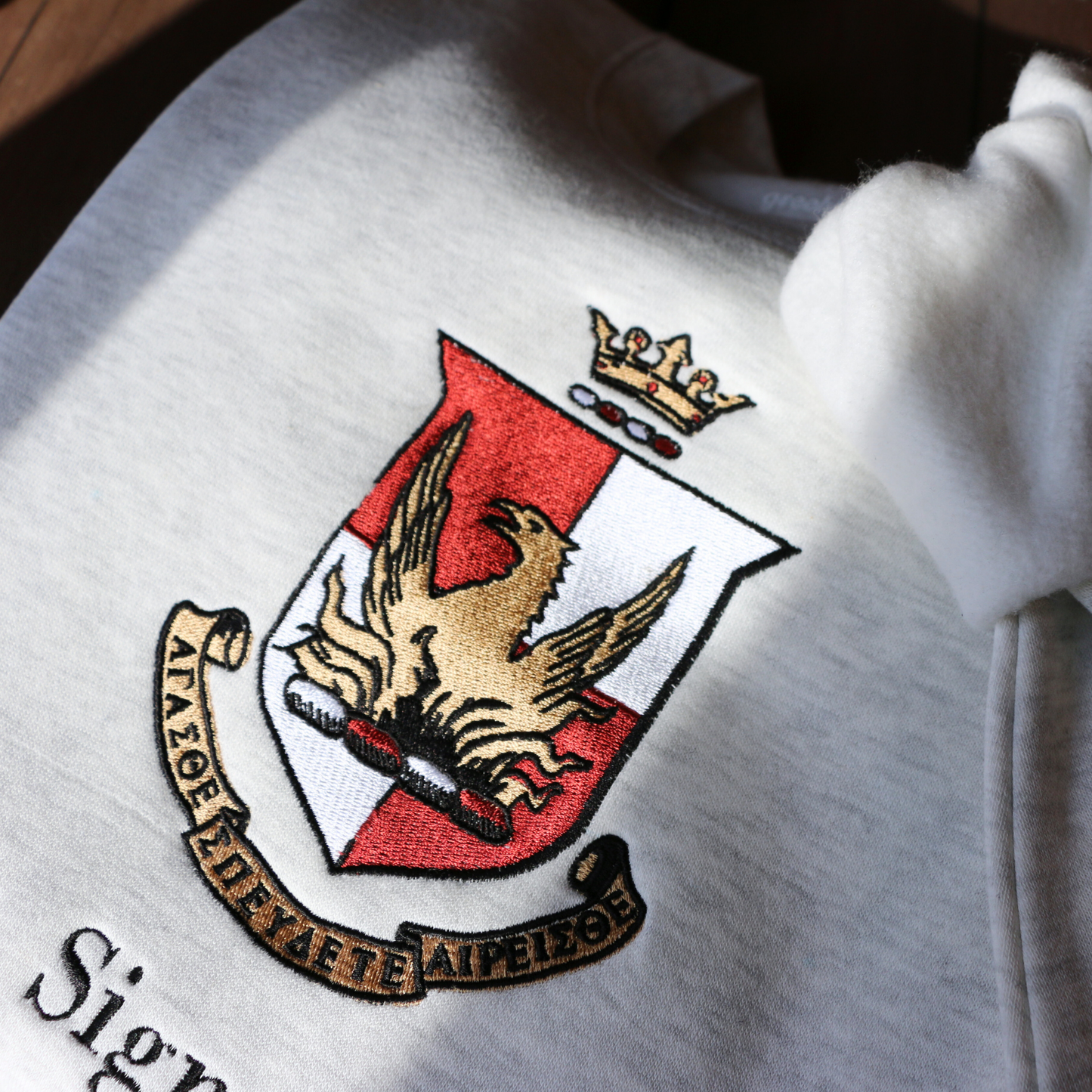 Detailed view of a light gray crewneck sweatshirt laid flat on a wooden surface, showcasing the Alpha Sigma Alpha sorority crest embroidered in the center. The crest displays a red shield with a golden phoenix, topped by a crown, and surrounded by a scroll bearing the sorority's Greek motto and name.