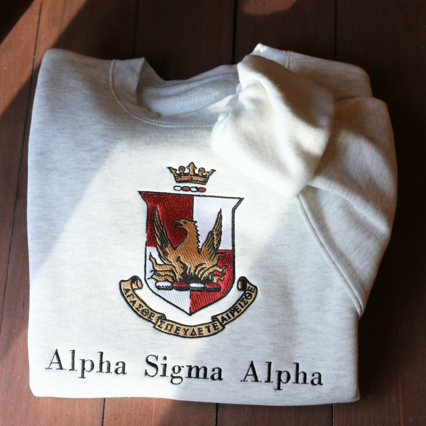 Light gray crewneck sweatshirt laid flat on a wooden surface, showcasing the Alpha Sigma Alpha sorority crest embroidered in the center. The crest displays a red shield with a golden phoenix, topped by a crown, and surrounded by a scroll bearing the sorority's Greek motto and name.