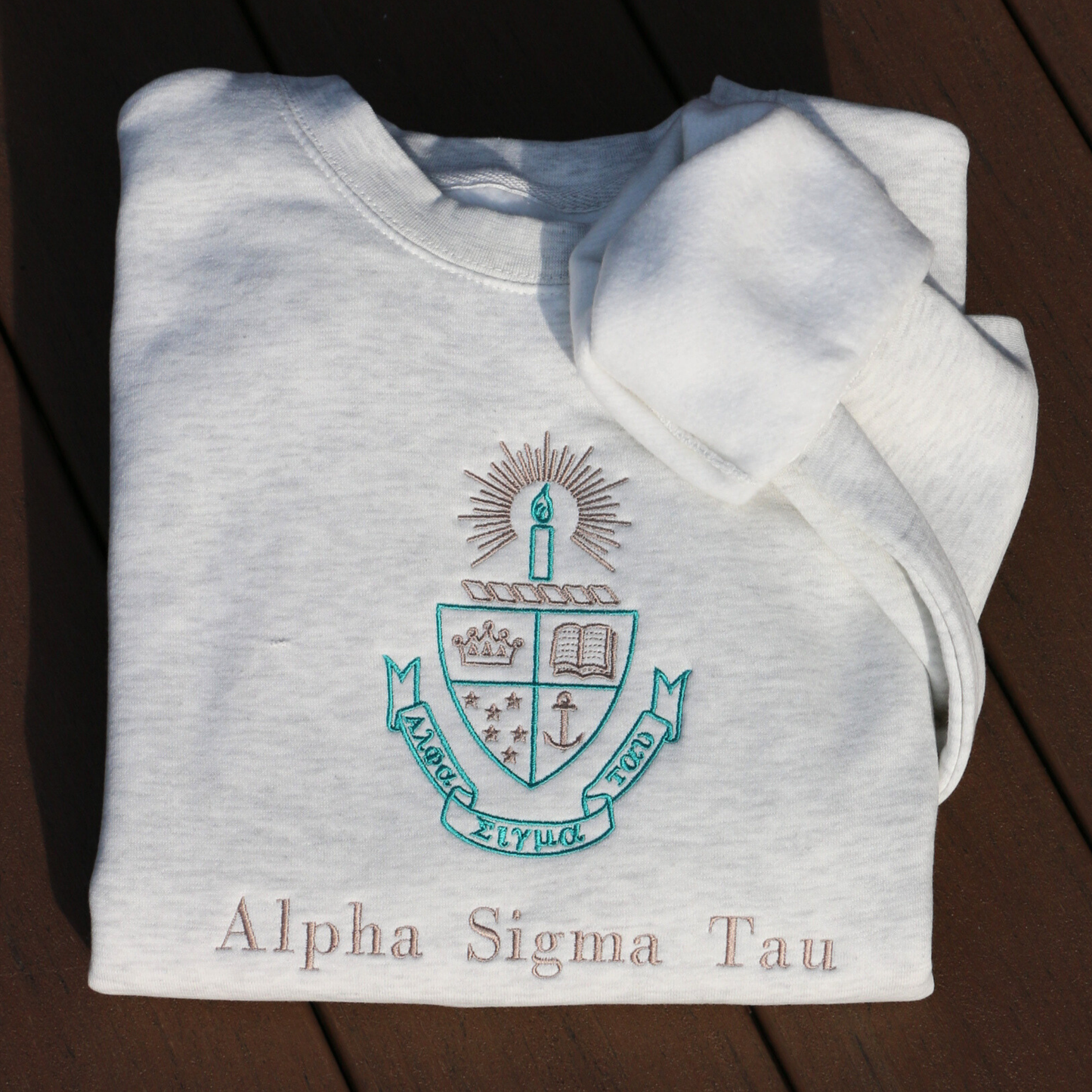 Light gray crewneck sweatshirt laid flat on a dark wood surface, featuring the embroidered crest of Alpha Sigma Tau. The crest includes a shield divided into four, showcasing a book, a crown, an anchor, and stars in teal and gold, with a candle and a golden light above. Below, the sorority name 'Alpha Sigma Tau' is embroidered below in gold.