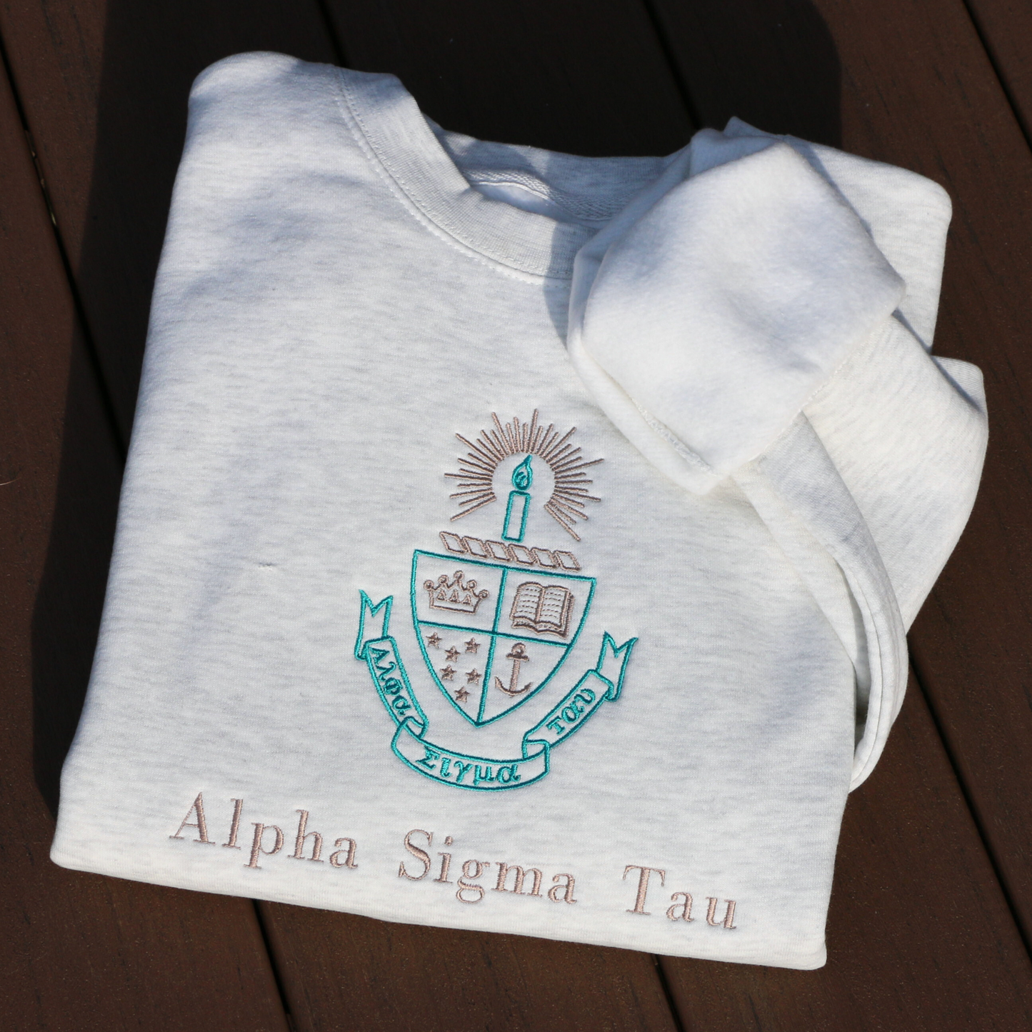 Light gray crewneck sweatshirt laid flat on a dark wood surface, featuring the embroidered crest of Alpha Sigma Tau. The crest includes a shield divided into four, showcasing a book, a crown, an anchor, and stars in teal and gold, with a candle and a golden light above. Below, the sorority name 'Alpha Sigma Tau' is embroidered below in gold.