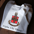 Load image into Gallery viewer, Gray crewneck sweatshirt laid flat on a dark wooden surface, featuring a large, detailed embroidery of the Alpha Chi Omega sorority crest in the center. The crest includes a red shield a gold book and stars. The sorority's name is embroidered below in black.