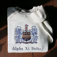 Load image into Gallery viewer, Light gray crewneck sweatshirt laid flat on a wooden surface, showcasing an elaborate embroidered crest of Alpha Xi Delta. The crest features two blue griffins flanking a shield topped by a head of armor and a pink flower. Below, the sorority name 'Alpha Xi Delta' is embroidered.