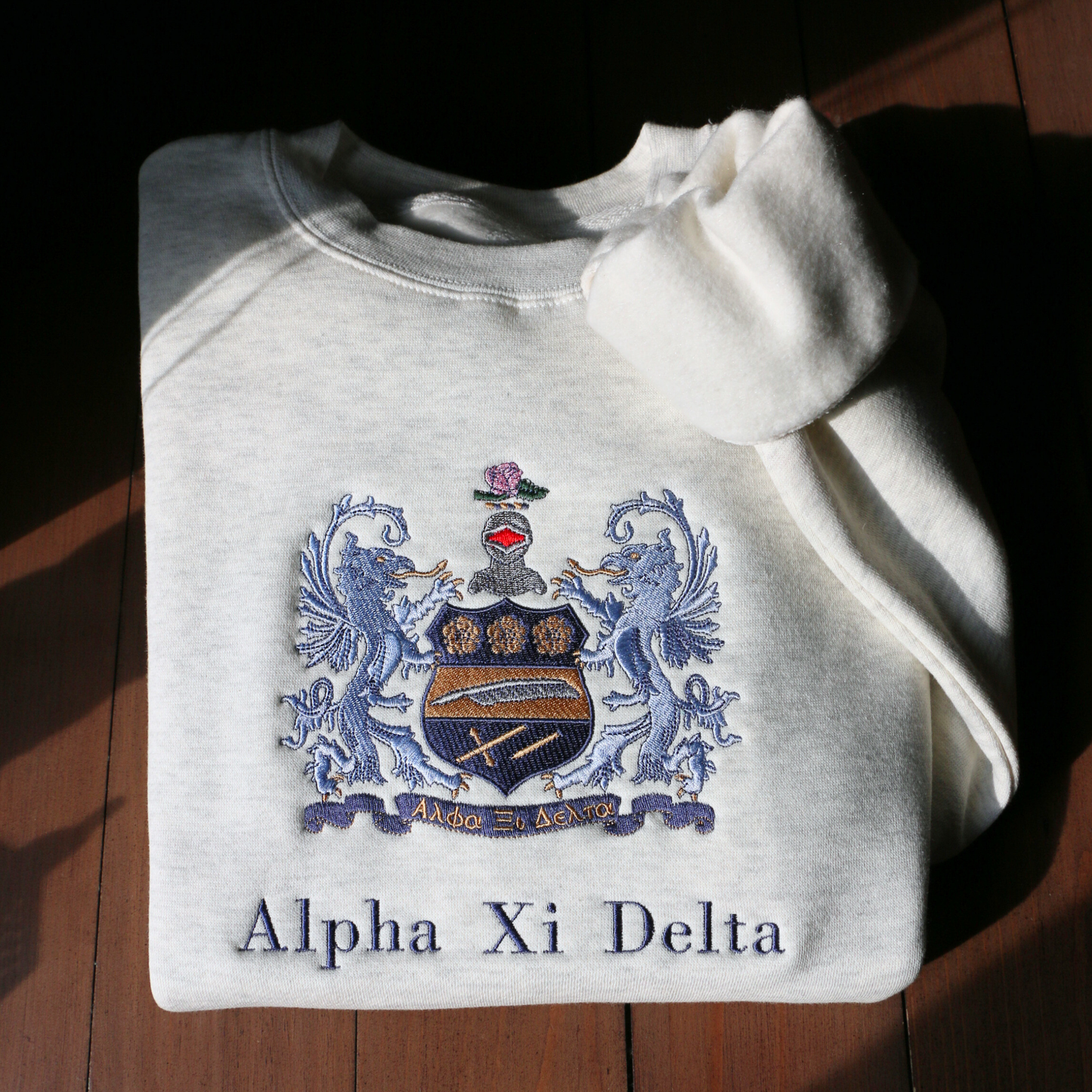 Light gray crewneck sweatshirt laid flat on a wooden surface, showcasing an elaborate embroidered crest of Alpha Xi Delta. The crest features two blue griffins flanking a shield topped by a head of armor and a pink flower. Below, the sorority name 'Alpha Xi Delta' is embroidered.