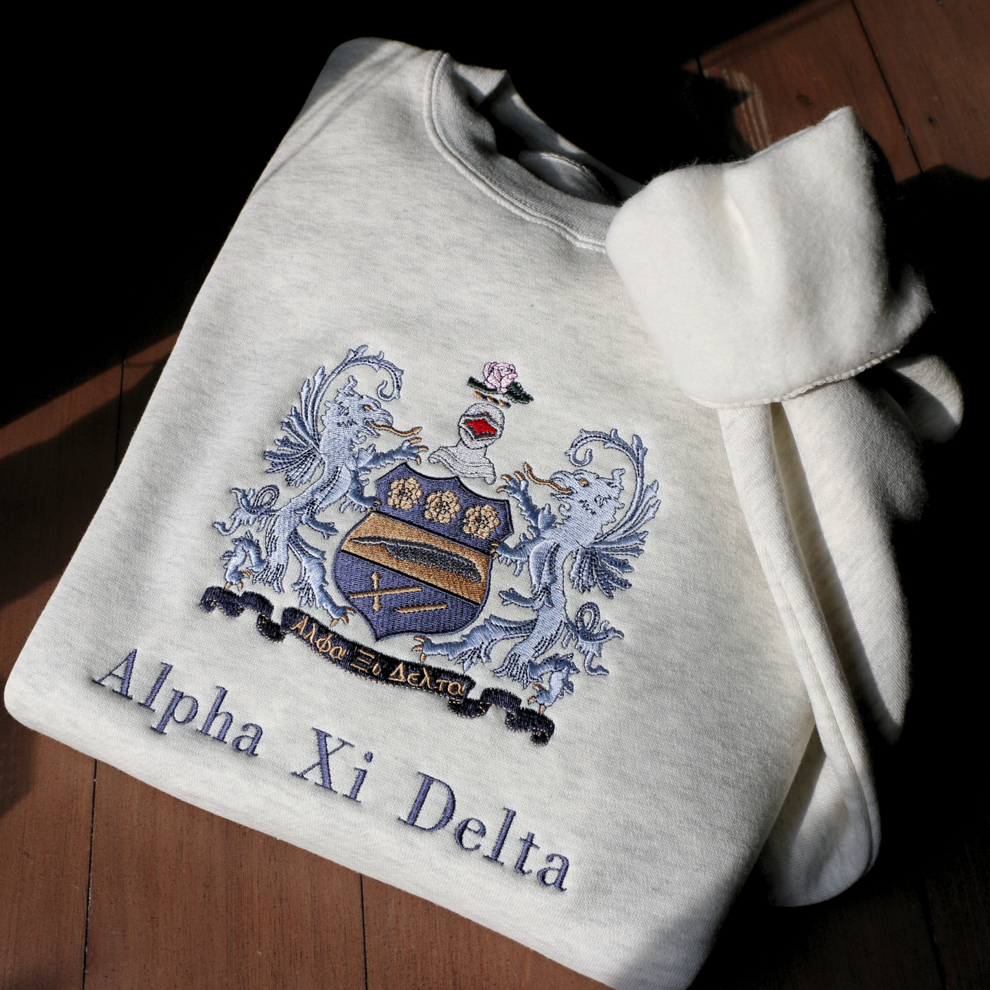 Light gray crewneck sweatshirt laid flat on a wooden surface, showcasing an elaborate embroidered crest of Alpha Xi Delta. The crest features two blue griffins flanking a shield topped by a head of armor and a pink flower. Below, the sorority name 'Alpha Xi Delta' is embroidered.