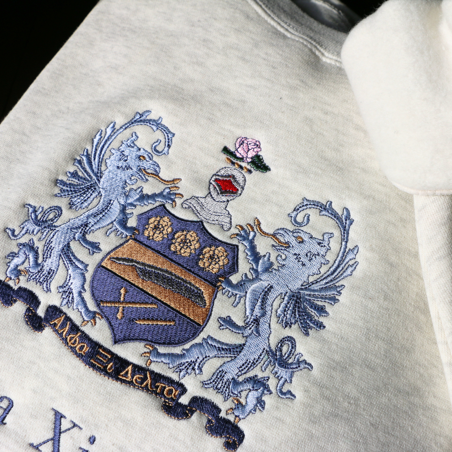 Detailed view of a light gray crewneck sweatshirt laid flat on a wooden surface, showcasing an elaborate embroidered crest of Alpha Xi Delta. The crest features two blue griffins flanking a shield topped by a head of armor and a pink flower. Below, the sorority name 'Alpha Xi Delta' is embroidered.