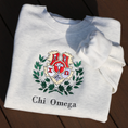 Load image into Gallery viewer, Light gray crewneck sweatshirt laid flat, featuring an embroidered crest of Chi Omega. The crest showcases a red and yellow shield adorned with a white skull and crossed bones, and an owl, surrounded by green leaves, and the sorority name 'Chi Omega' in elegant black lettering below.