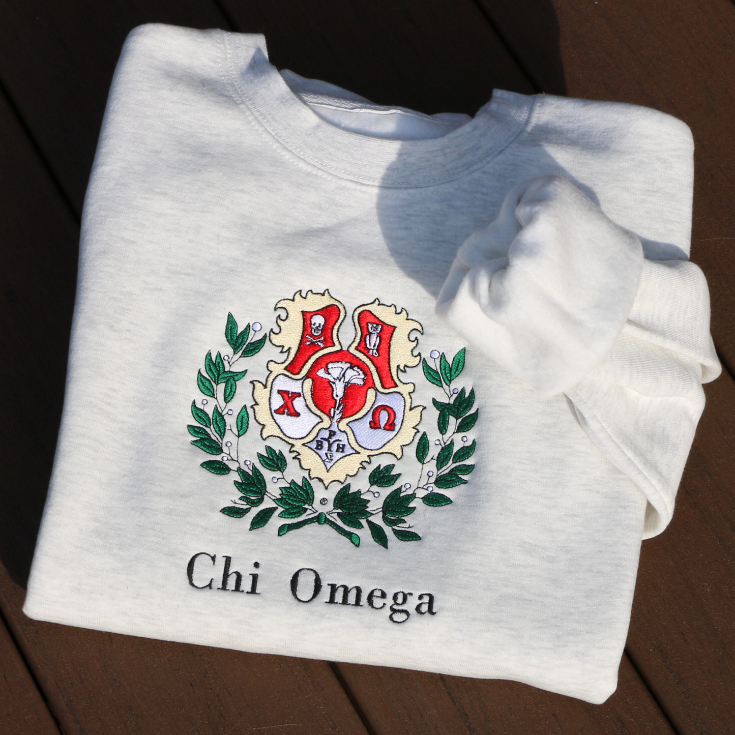 Light gray crewneck sweatshirt laid flat, featuring an embroidered crest of Chi Omega. The crest showcases a red and yellow shield adorned with a white skull and crossed bones, and an owl, surrounded by green leaves, and the sorority name 'Chi Omega' in elegant black lettering below.