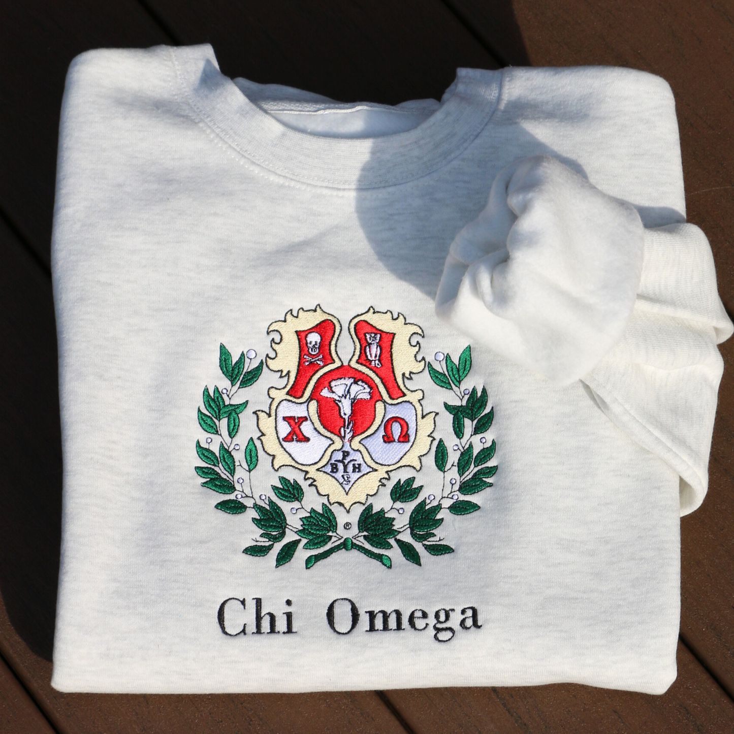 Light gray crewneck sweatshirt laid flat, featuring an embroidered crest of Chi Omega. The crest showcases a red and yellow shield adorned with a white skull and crossed bones, and an owl, surrounded by green leaves, and the sorority name 'Chi Omega' in elegant black lettering below.