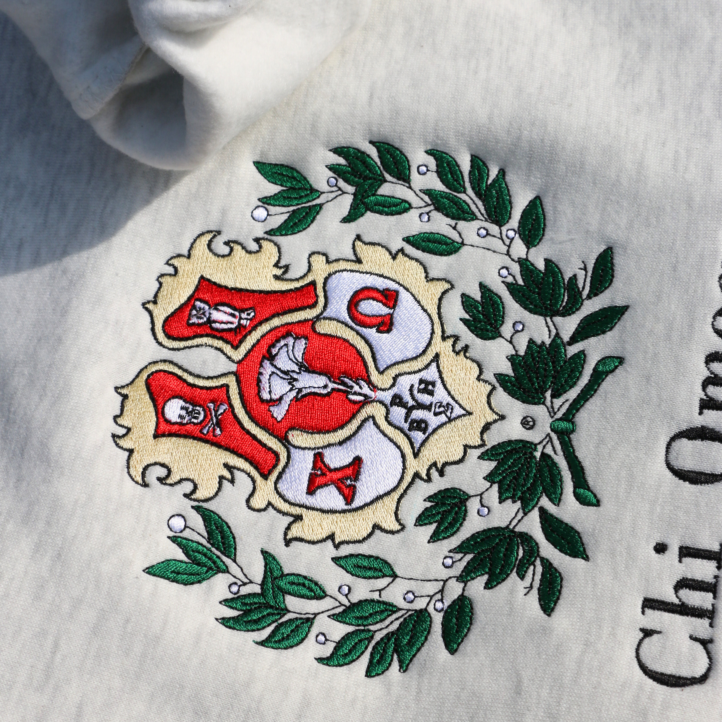 Detailed view of a light gray crewneck sweatshirt laid flat, featuring an embroidered crest of Chi Omega. The crest showcases a red and yellow shield adorned with a white skull and crossed bones, and an owl, surrounded by green leaves, and the sorority name 'Chi Omega' in elegant black lettering below.