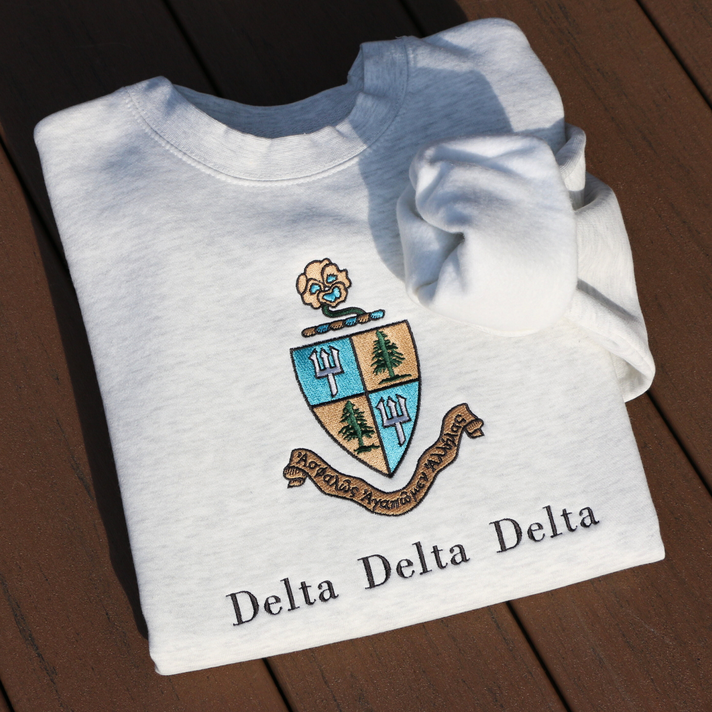 Light gray crewneck sweatshirt embroidered with Delta Delta Delta's crest, featuring a blue and gold shield with pine trees and tridents. The sorority’s Greek motto “Let us steadfastly love one another,” is inscribed in Greek on a scroll below the shield. Below this, the sorority's name is embroidered in black.