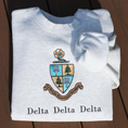 Load image into Gallery viewer, Light gray crewneck sweatshirt embroidered with Delta Delta Delta's crest, featuring a blue and gold shield with pine trees and tridents. The sorority’s Greek motto “Let us steadfastly love one another,” is inscribed in Greek on a scroll below the shield. Below this, the sorority's name is embroidered in black.