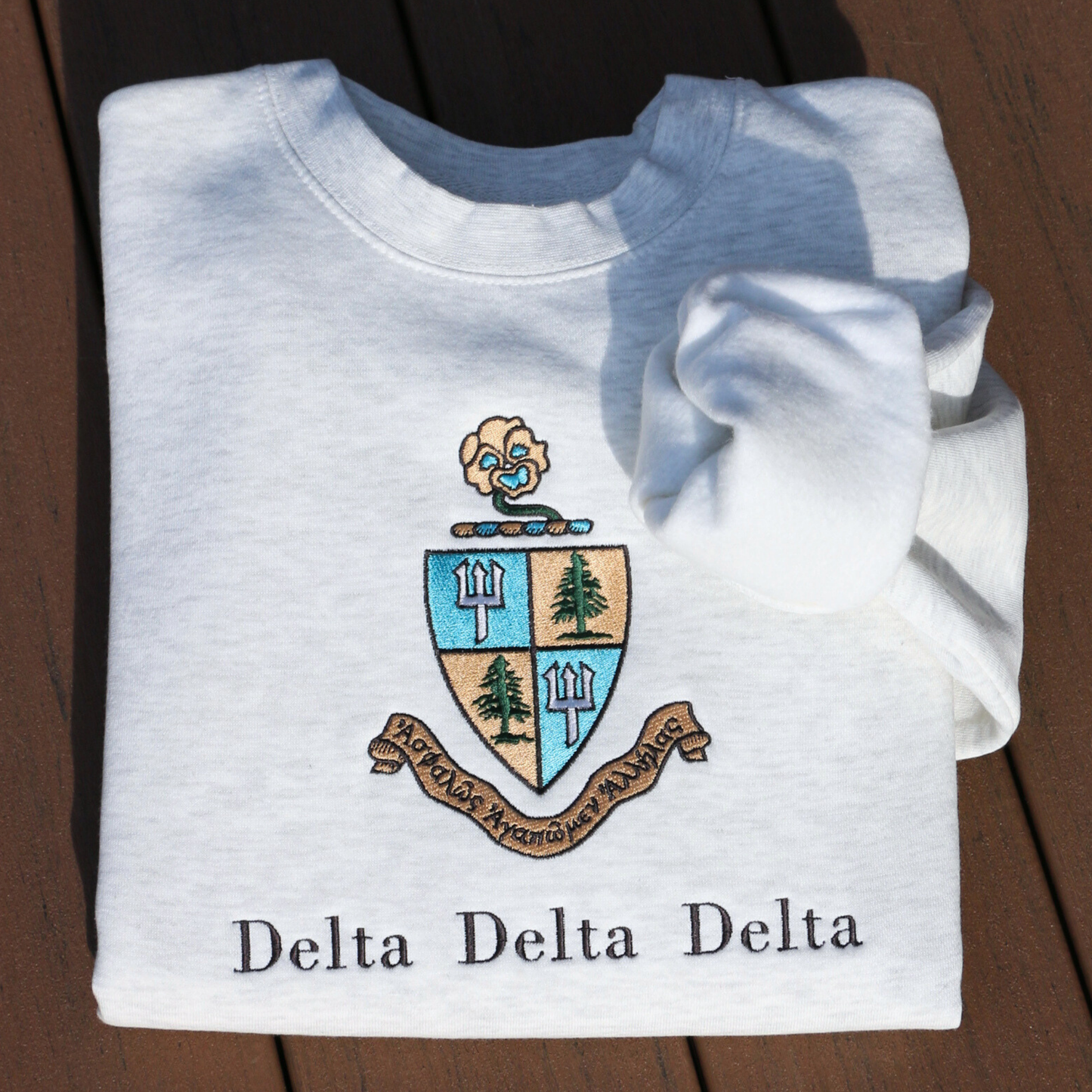 Light gray crewneck sweatshirt embroidered with Delta Delta Delta's crest, featuring a blue and gold shield with pine trees and tridents. The sorority’s Greek motto “Let us steadfastly love one another,” is inscribed in Greek on a scroll below the shield. Below this, the sorority's name is embroidered in black.