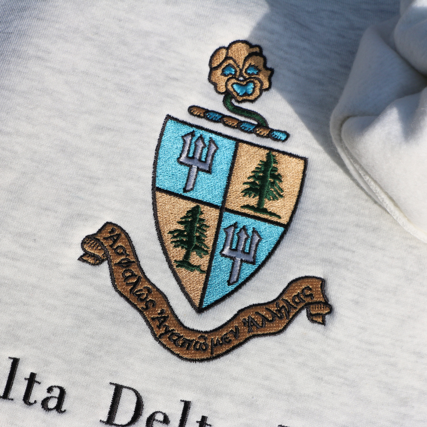 Detailed view of a light gray crewneck sweatshirt embroidered with Delta Delta Delta's crest, featuring a blue and gold shield with pine trees and tridents. The sorority’s Greek motto “Let us steadfastly love one another,” is inscribed in Greek on a scroll below the shield. Below this, the sorority's name is embroidered in black.