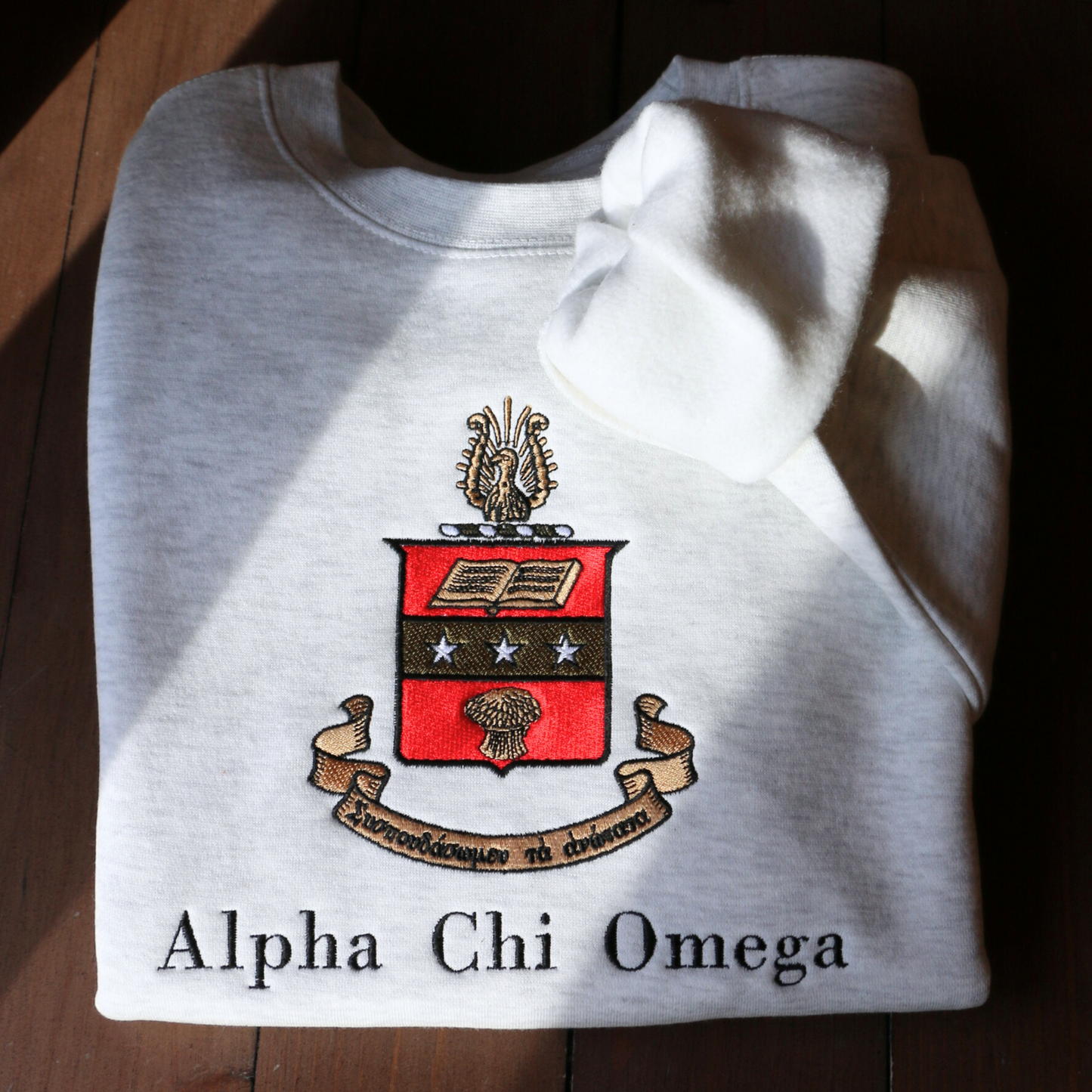 Gray crewneck sweatshirt laid flat on a dark wooden surface, featuring a large, detailed embroidery of the Alpha Chi Omega sorority crest in the center. The crest includes a red shield a gold book and stars. The sorority's name is embroidered below in black.