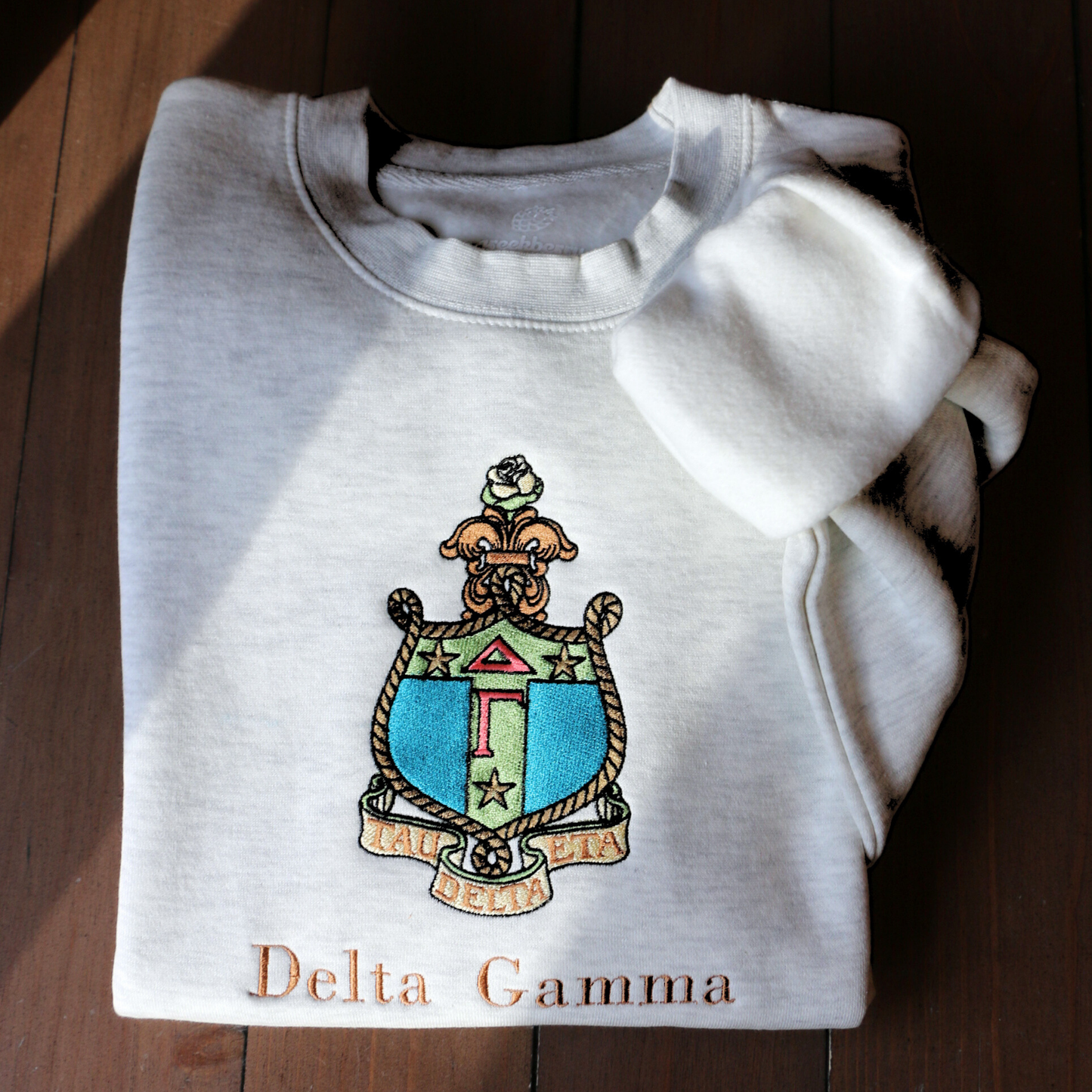 Delta Gamma's crest embroidered on a light gray sweatshirt, featuring a rose, stars, rope, and the greek letters. Below, 'Delta Gamma' is written in an elegant brown script.