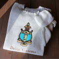 Load image into Gallery viewer, Delta Gamma's crest embroidered on a light gray sweatshirt, featuring a rose, stars, rope, and the greek letters. Below, 'Delta Gamma' is written in an elegant brown script.