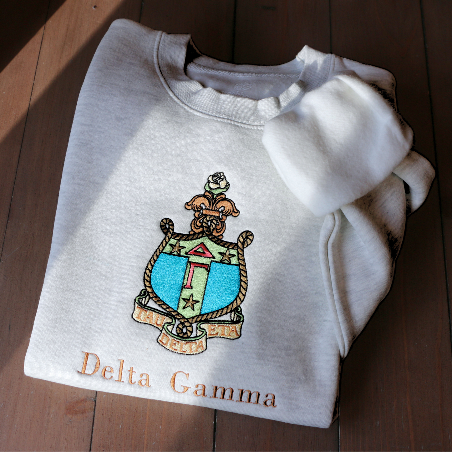 Delta Gamma's crest embroidered on a light gray sweatshirt, featuring a rose, stars, rope, and the greek letters. Below, 'Delta Gamma' is written in an elegant brown script.