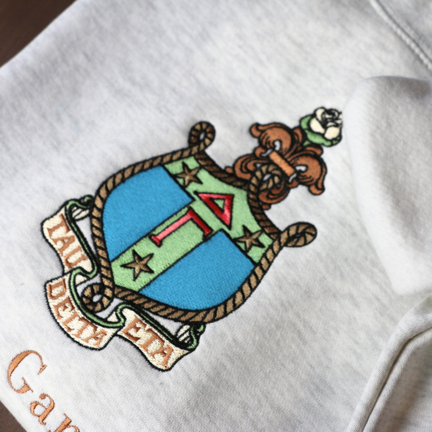 Detailed view of Delta Gamma's crest embroidered on a light gray sweatshirt, featuring a rose, stars, rope, and the greek letters. Below, 'Delta Gamma' is written in an elegant brown script.