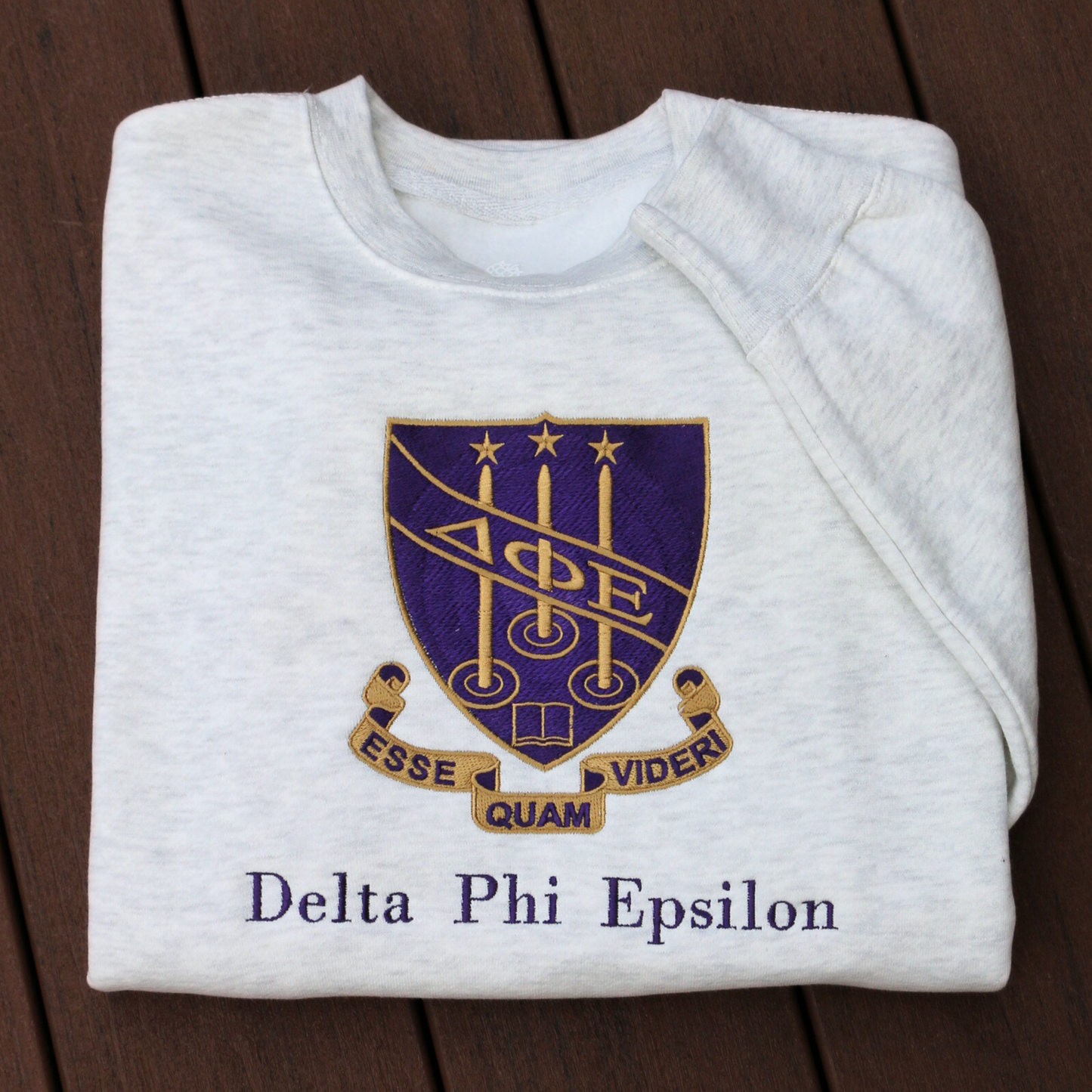 Delta Phi Epsilon's crest embroidered on a light gray sweatshirt, featuring a purple shield and the sorority's letters. The crest is surrounded by a scroll inscribed with the Latin motto 'Esse Quam Videri' (To be, rather than to seem). Below this, the sorority's name is embroidered in a dark purple.