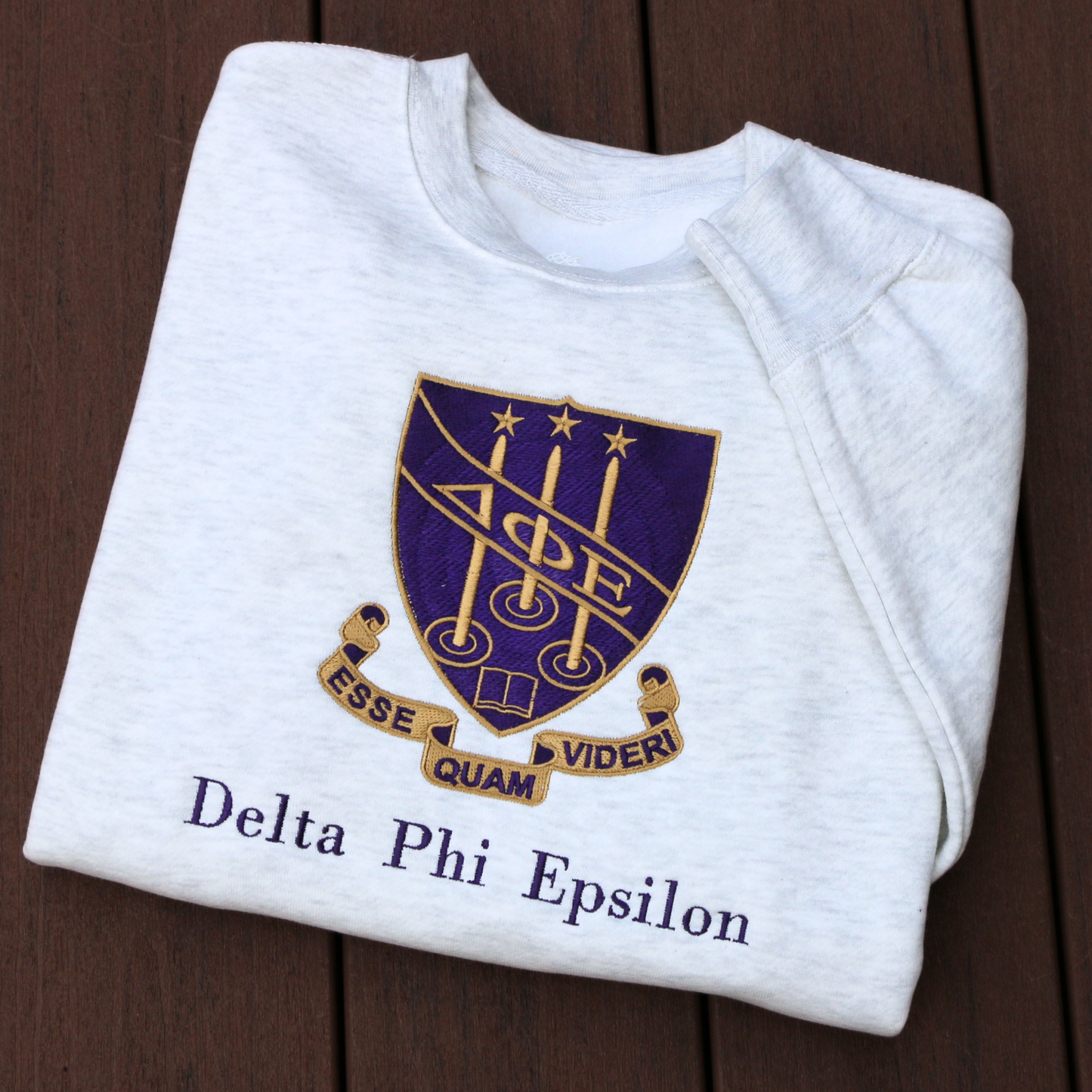 Delta Phi Epsilon's crest embroidered on a light gray sweatshirt, featuring a purple shield and the sorority's letters. The crest is surrounded by a scroll inscribed with the Latin motto 'Esse Quam Videri' (To be, rather than to seem). Below this, the sorority's name is embroidered in a dark purple.