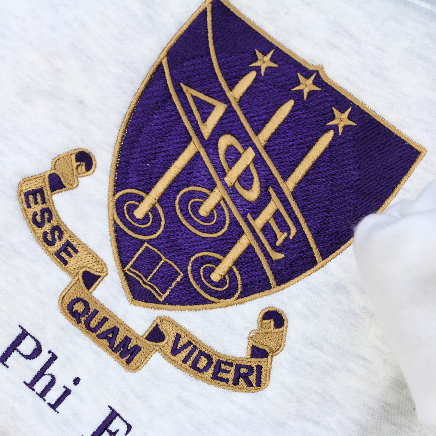 Detailed view of Delta Phi Epsilon's crest embroidered on a light gray sweatshirt, featuring a purple shield and the sorority's letters. The crest is surrounded by a scroll inscribed with the Latin motto 'Esse Quam Videri' (To be, rather than to seem). Below this, the sorority's name is embroidered in a dark purple.