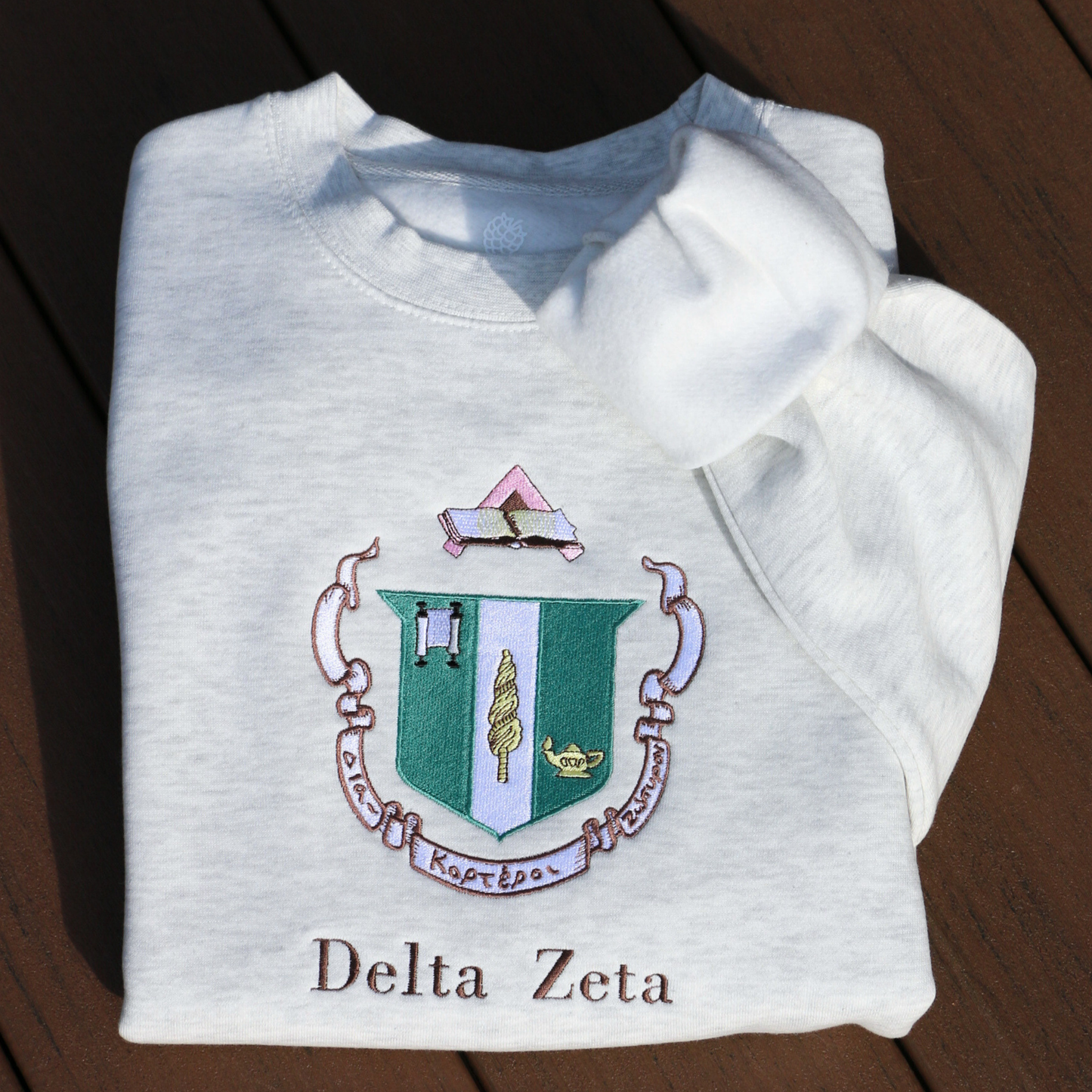 Delta Zeta's crest embroidered on a light gray sweatshirt, featuring a green shield with a roman lamp and scroll. Below, the sorority's name is embroidered in brown.
