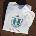 Load image into Gallery viewer, Delta Zeta's crest embroidered on a light gray sweatshirt, featuring a green shield with a roman lamp and scroll. Below, the sorority's name is embroidered in brown.