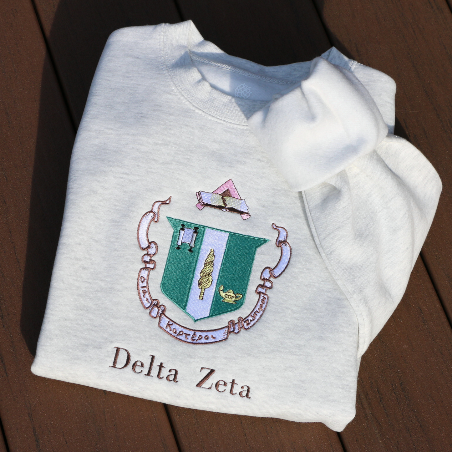 Delta Zeta's crest embroidered on a light gray sweatshirt, featuring a green shield with a roman lamp and scroll. Below, the sorority's name is embroidered in brown.