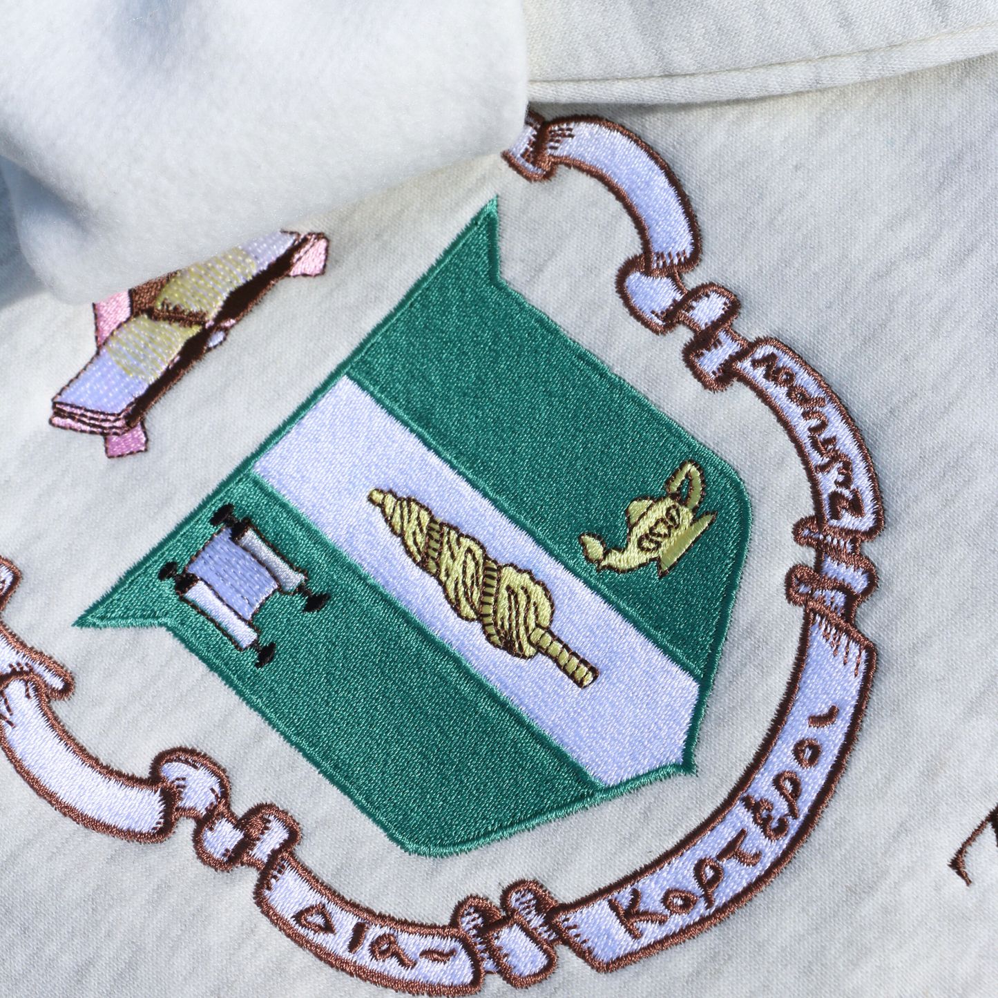 Detailed view of Delta Zeta's crest embroidered on a light gray sweatshirt, featuring a green shield with a roman lamp and scroll. Below, the sorority's name is embroidered in brown.