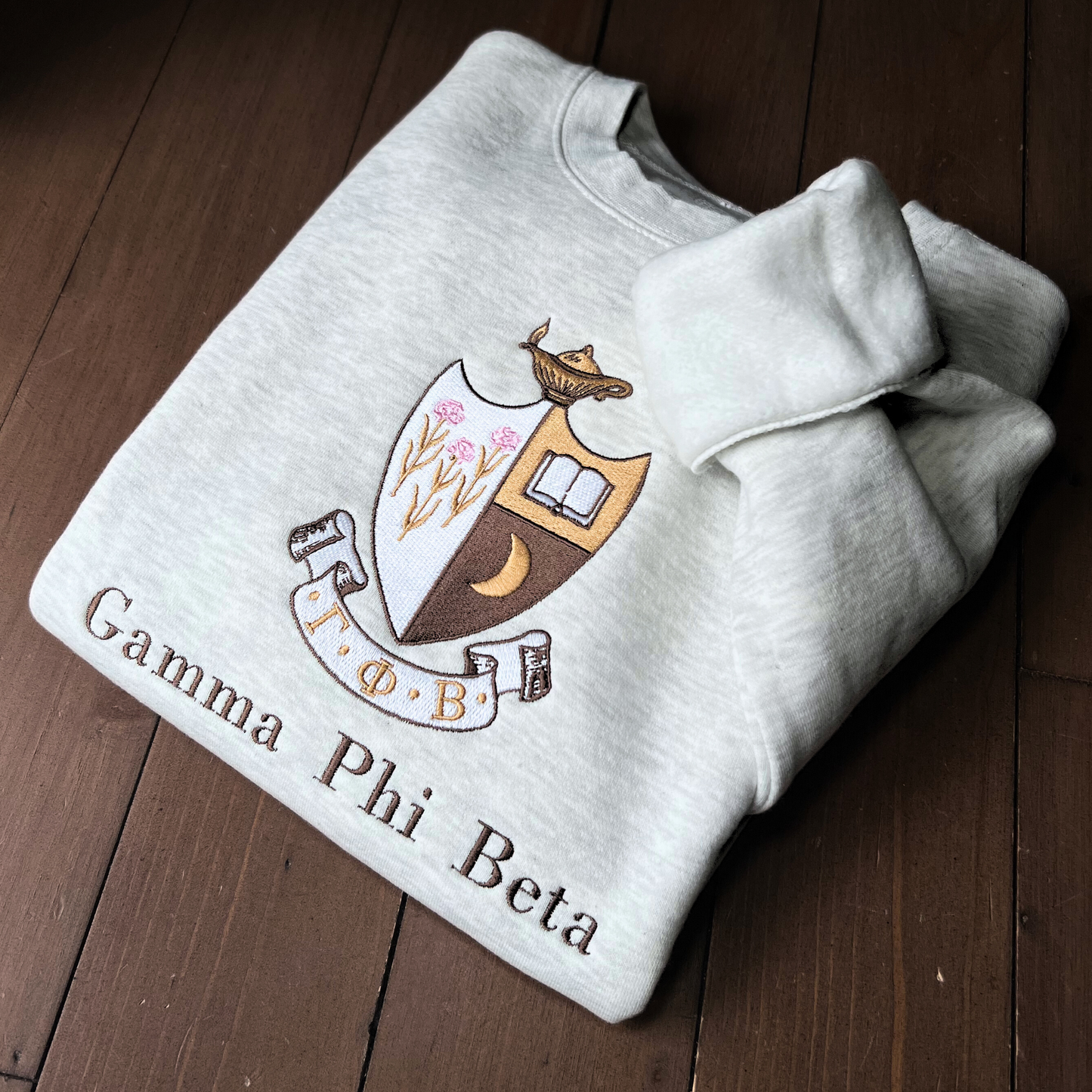 Gamma Phi Beta's crest embroidered on a light gray sweatshirt, featuring a brown shield with a golden crescent moon and pink flowers, and an open book, topped by a golden lamp, with 'Gamma Phi Beta' written below in elegant script.