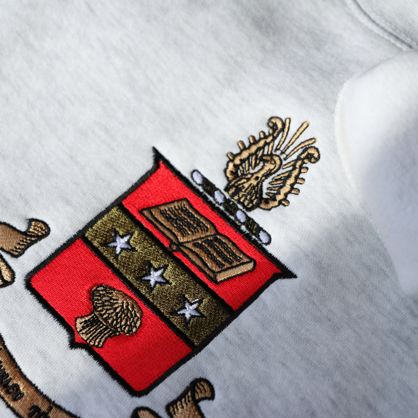 Detailed view of a gray crewneck sweatshirt laid flat on a dark wooden surface, featuring a large, detailed embroidery of the Alpha Chi Omega sorority crest in the center. The crest includes a red shield a gold book and stars. The sorority's name is embroidered below in black.