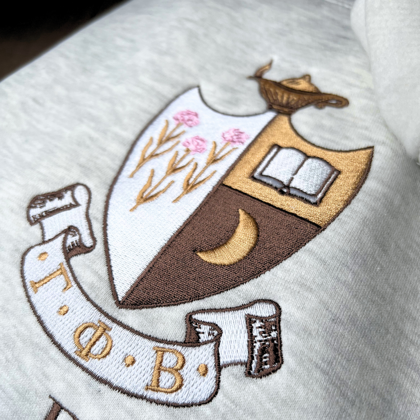 A detailed view of Gamma Phi Beta's crest embroidered on a light gray sweatshirt, featuring a brown shield with a golden crescent moon and pink flowers, and an open book, topped by a golden lamp, with 'Gamma Phi Beta' written below in elegant script.