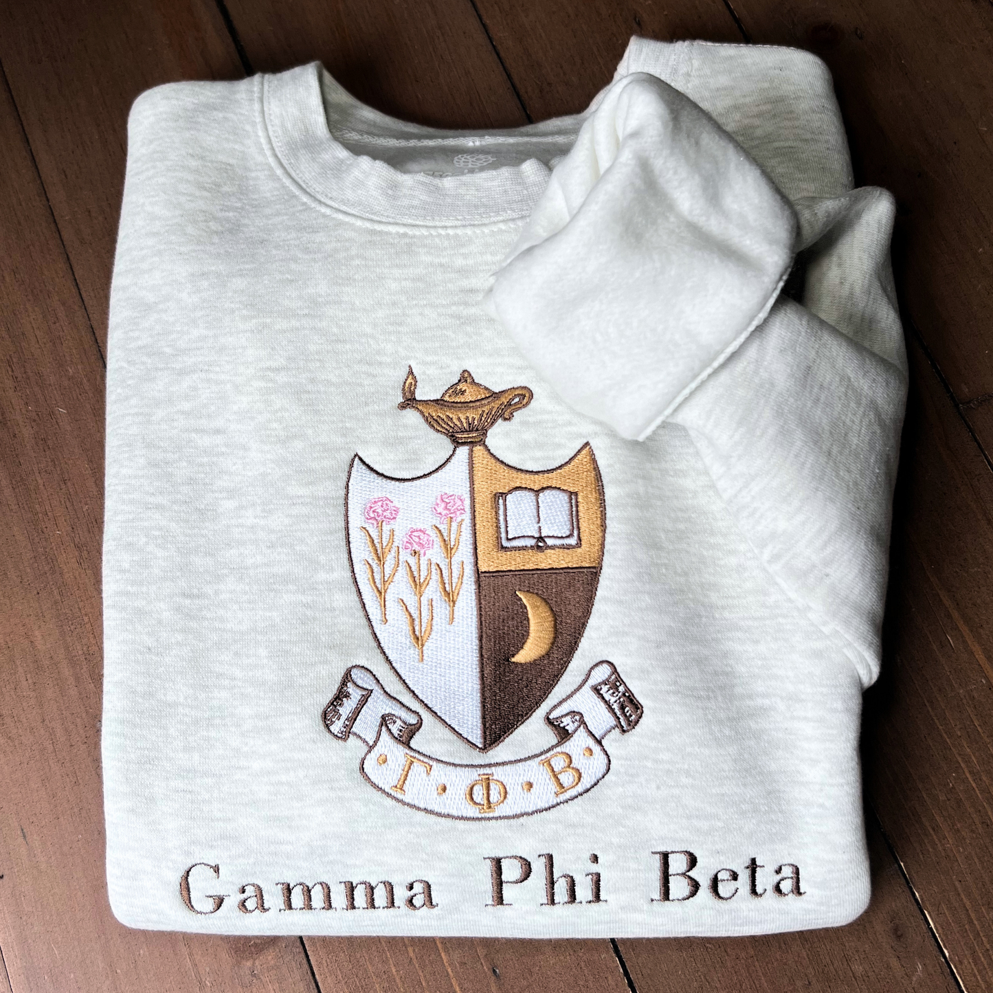 Gamma Phi Beta's crest embroidered on a light gray sweatshirt, featuring a brown shield with a golden crescent moon and pink flowers, and an open book, topped by a golden lamp, with 'Gamma Phi Beta' written below in elegant script.