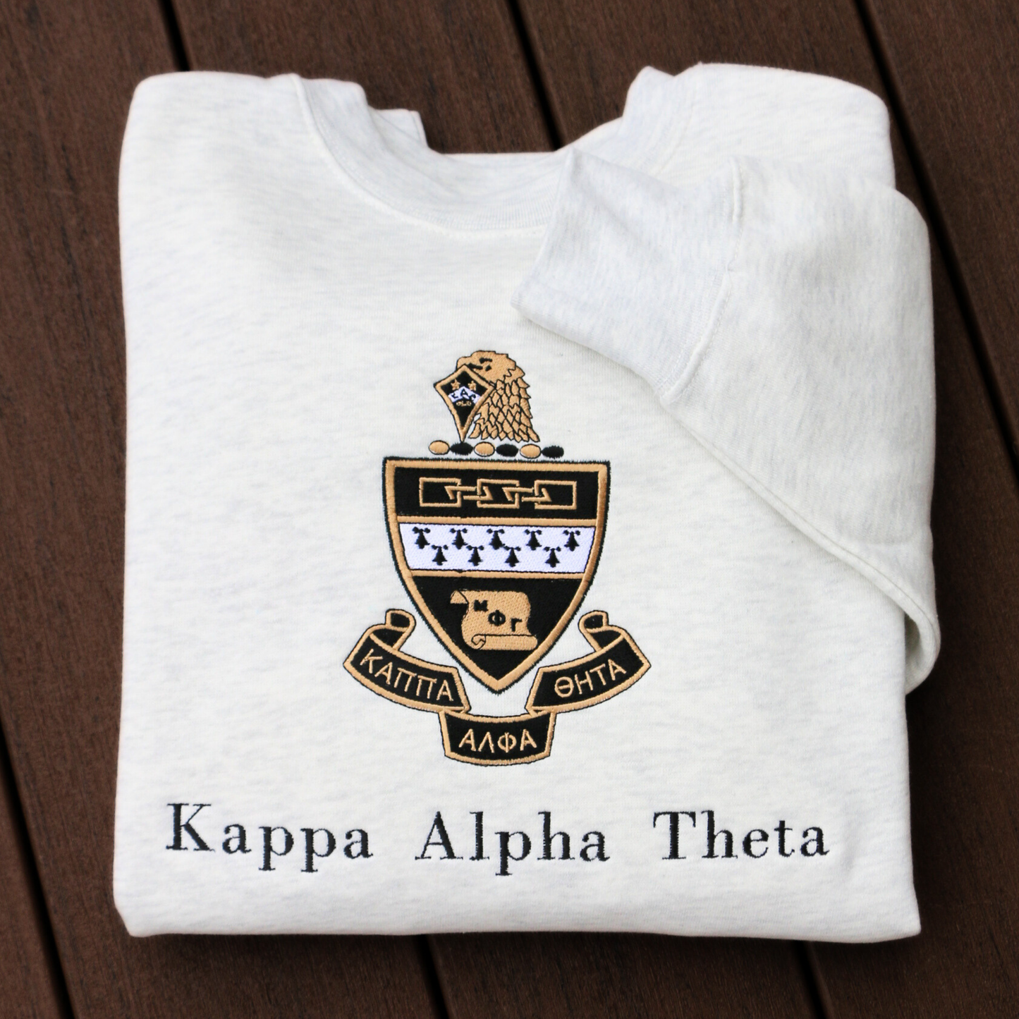 Kappa Alpha Theta's crest embroidered on a light gray sweatshirt, featuring a black and gold shield with white chevron, and twin stars, flanked by scrolls reading 'Kappa Alpha Theta' in black lettering.