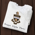 Load image into Gallery viewer, Kappa Alpha Theta's crest embroidered on a light gray sweatshirt, featuring a black and gold shield with white chevron, and twin stars, flanked by scrolls reading 'Kappa Alpha Theta' in black lettering.