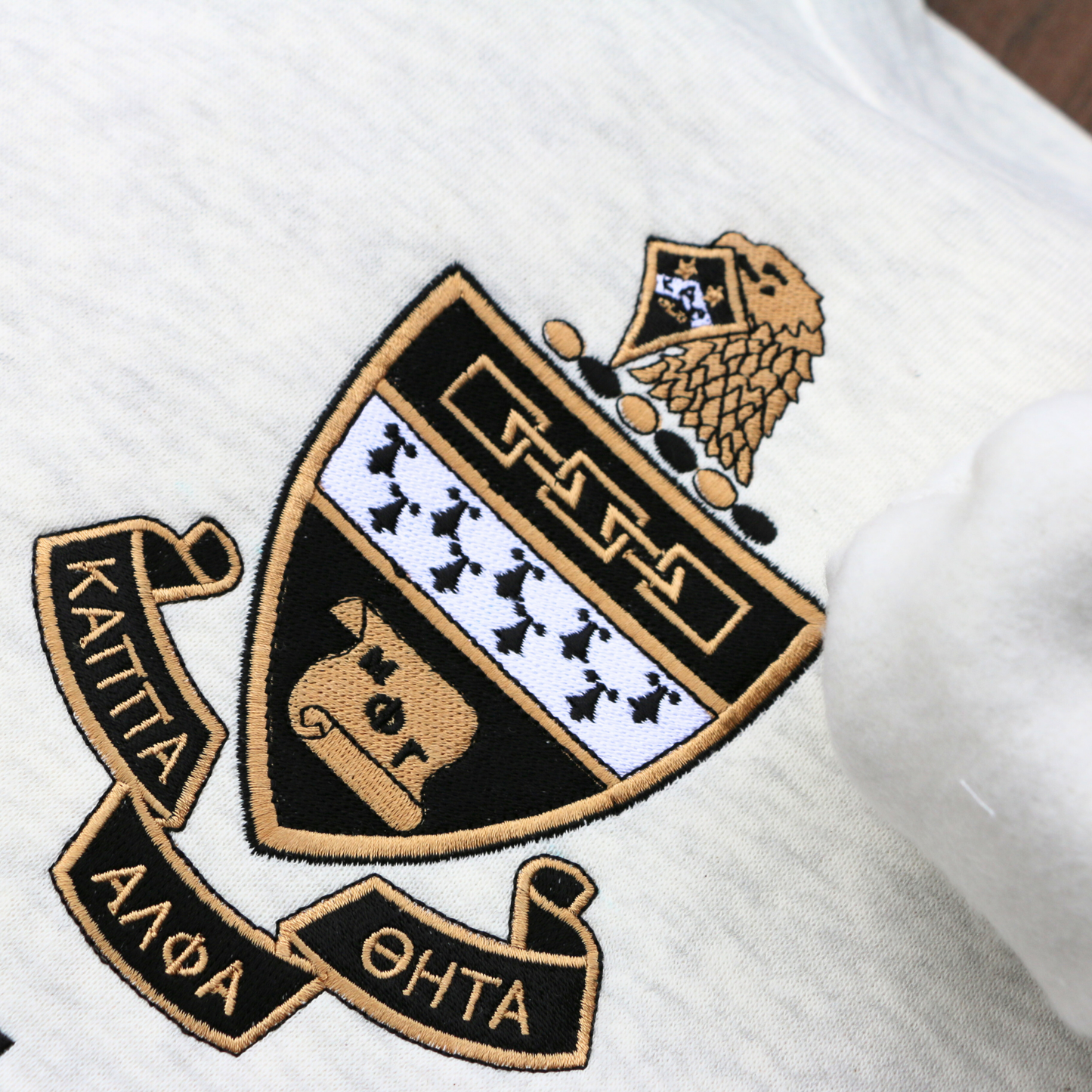 Detailed view of Kappa Alpha Theta's crest embroidered on a light gray sweatshirt, featuring a black and gold shield with white chevron, and twin stars, flanked by scrolls reading 'Kappa Alpha Theta' in black lettering.
