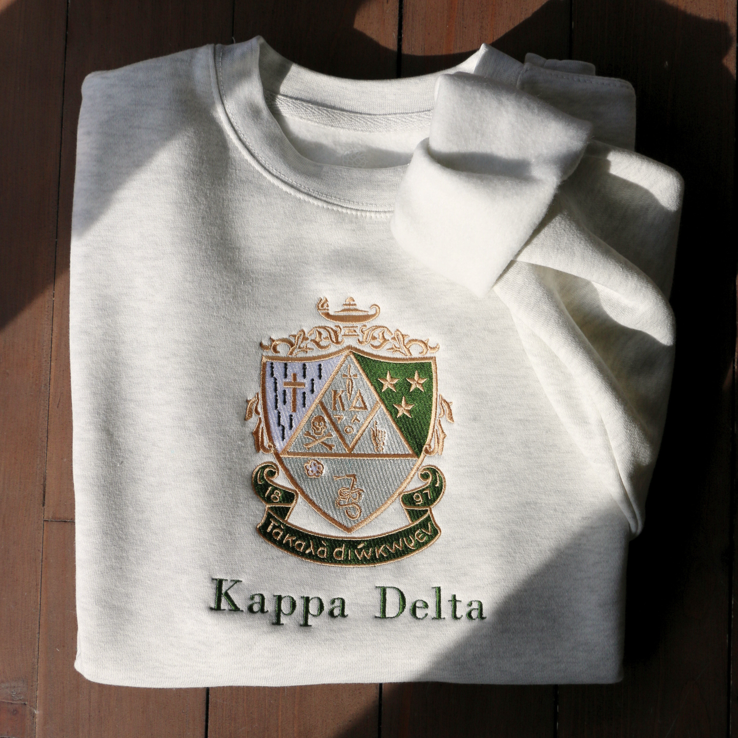 Kappa Delta's crest embroidered on a light gray sweatshirt, featuring a green and gold shield with stars, a skull and crossbones, a cross, the sorority's greek letters, a lamp, and a scroll. a white rose and stars, underlined by a scroll with 'Τa Kala Diokomen' written on it. Below, the sorority name is embroidered in dark green.