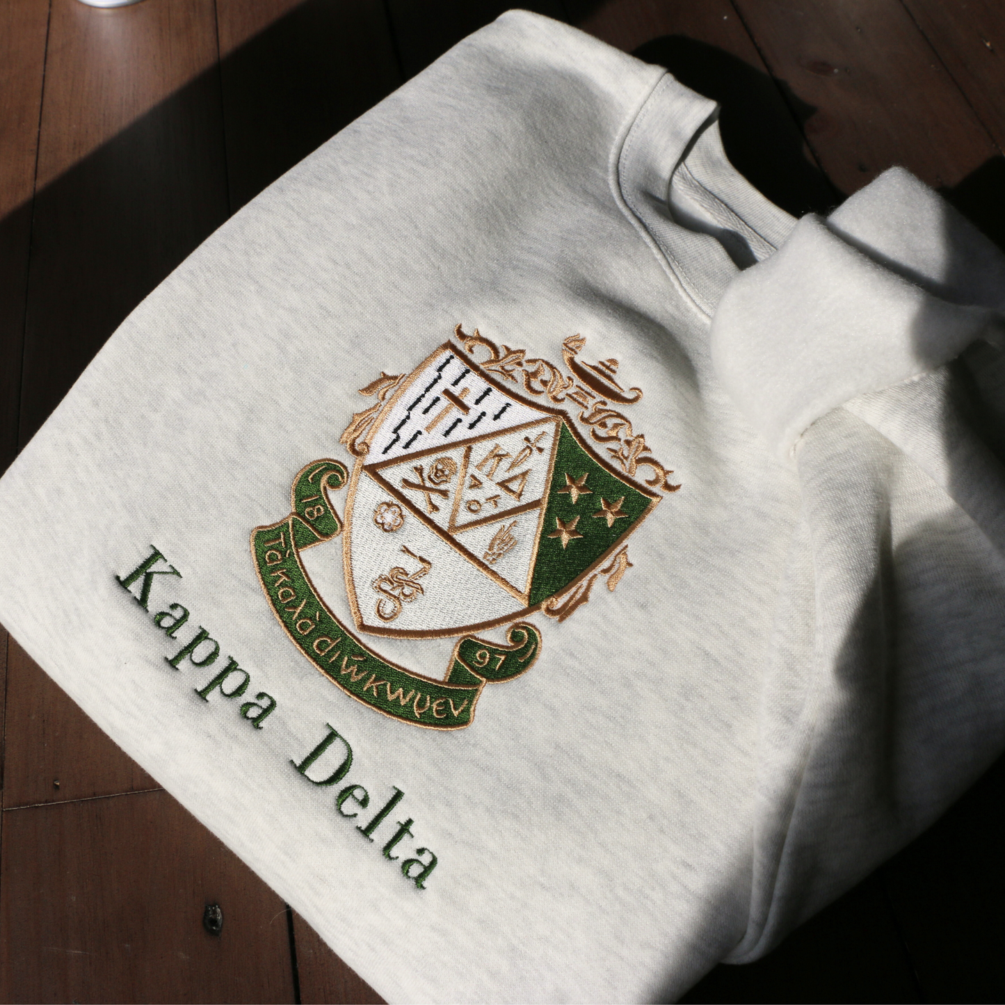 Kappa Delta's crest embroidered on a light gray sweatshirt, featuring a green and gold shield with stars, a skull and crossbones, a cross, the sorority's greek letters, a lamp, and a scroll. a white rose and stars, underlined by a scroll with 'Τa Kala Diokomen' written on it. Below, the sorority name is embroidered in dark green.