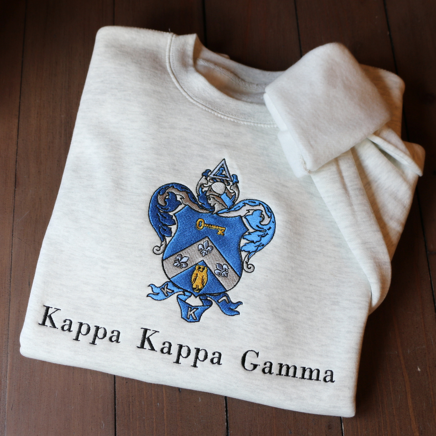 Kappa Kappa Gamma's crest embroidered on a light gray sweatshirt, featuring a blue shield with a golden key and fleur-de-lis, with the sorority name 'Kappa Kappa Gamma' in black lettering beneath.