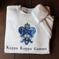 Load image into Gallery viewer, Kappa Kappa Gamma's crest embroidered on a light gray sweatshirt, featuring a blue shield with a golden key and fleur-de-lis, with the sorority name 'Kappa Kappa Gamma' in black lettering beneath.