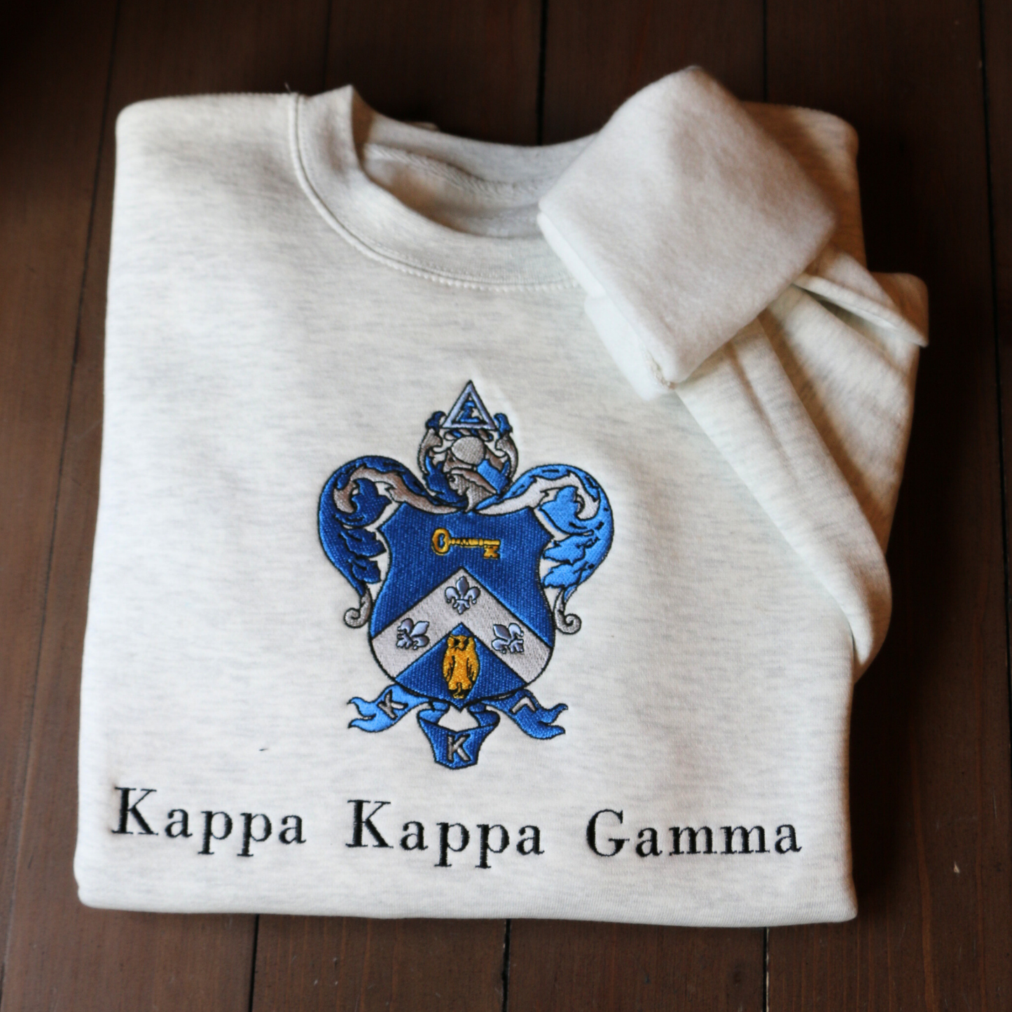 Kappa Kappa Gamma's crest embroidered on a light gray sweatshirt, featuring a blue shield with a golden key and fleur-de-lis, with the sorority name 'Kappa Kappa Gamma' in black lettering beneath.