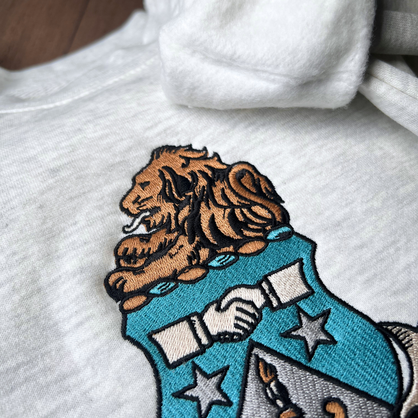 Detailed view of a gray crewneck sweatshirt featuring the Alpha Delta Pi sorority crest embroidered in the center. The crest includes a brown lion atop a blue shield with white stars. Below, the sorority name is embroidered in black.