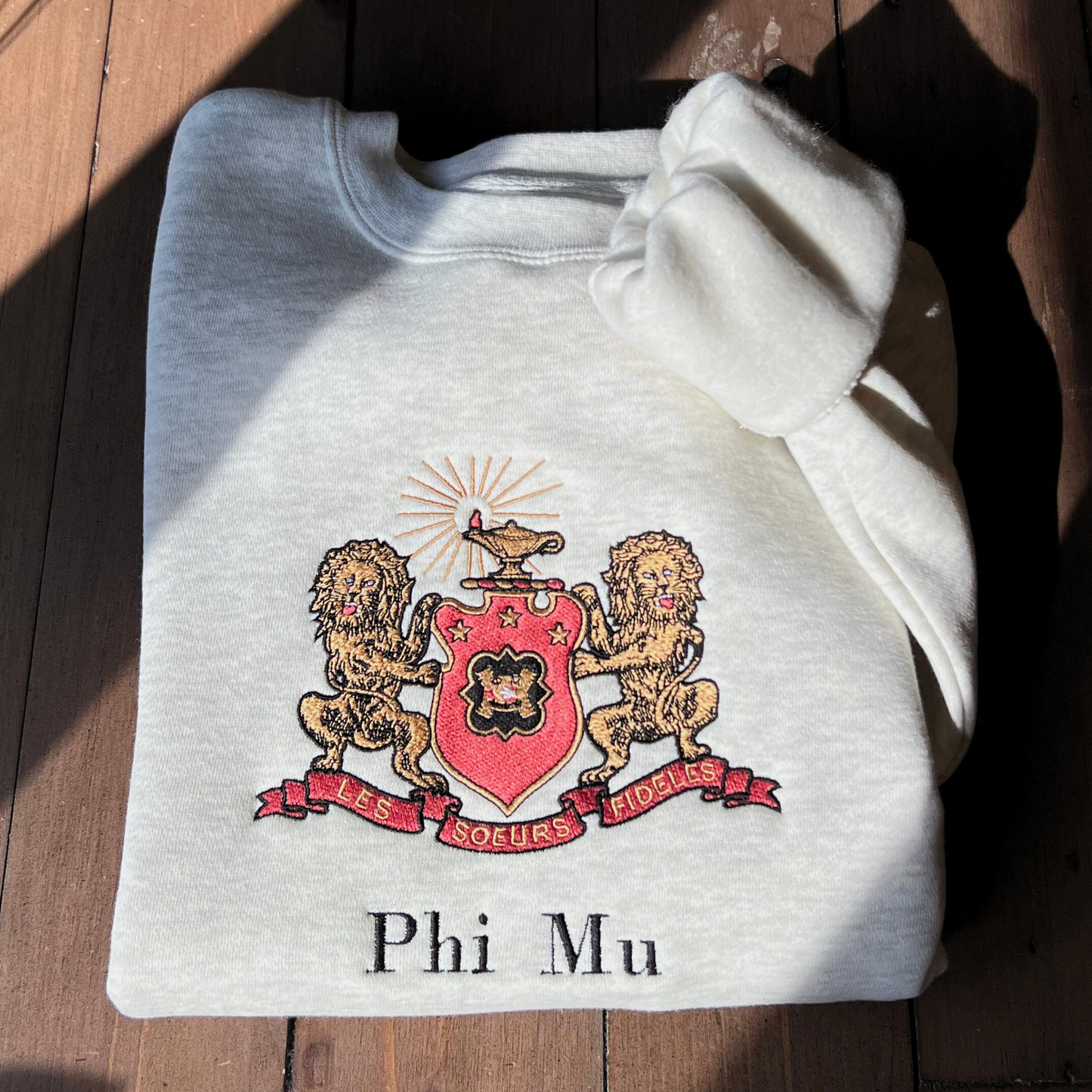 Phi Mu's crest embroidered on a light gray sweatshirt, featuring a shield guarded by gold lions. The shield includes the phi mu badge and under it is Phi Mu's open motto 'Les Soeurs Fideles' which translates to 'the Faithful Sisters'. Above the shield is the crest, the lighted lamp of knowledge in gold. Below the sorority name is embroidered in black.