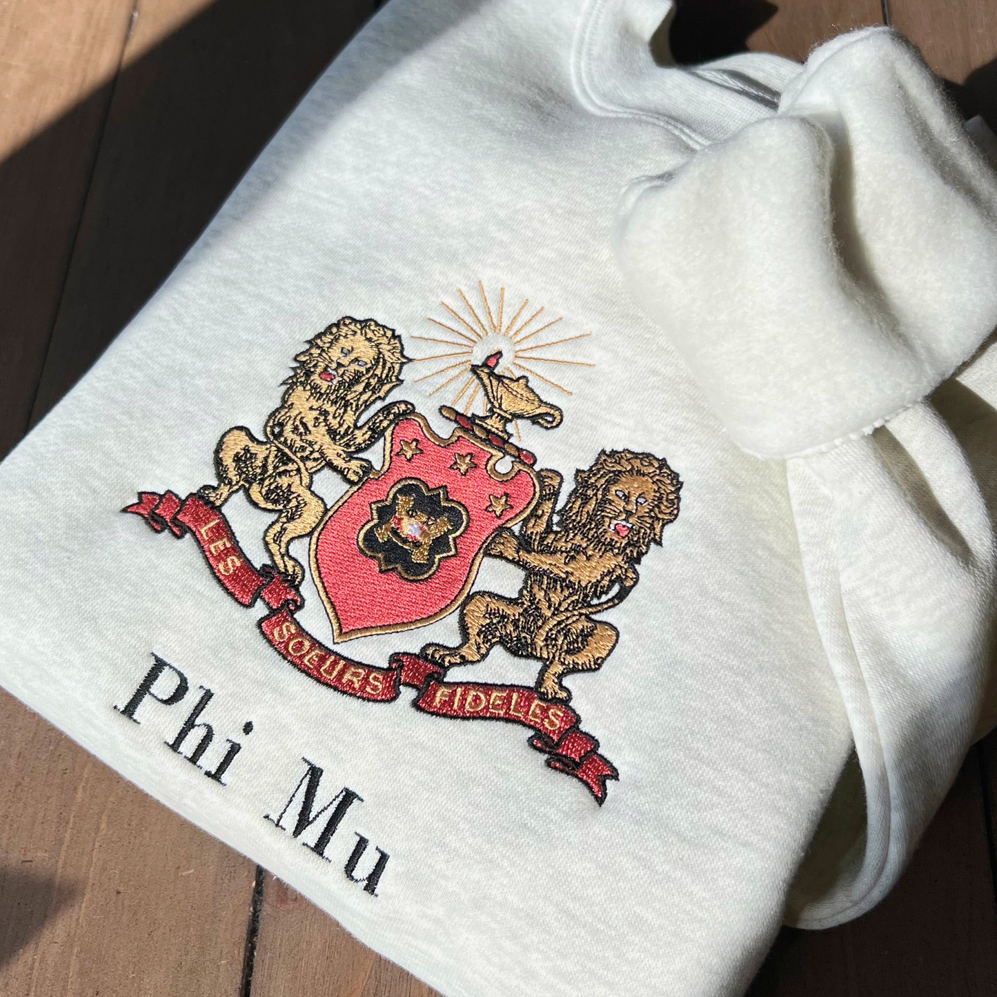 Phi Mu's crest embroidered on a light gray sweatshirt, featuring a shield guarded by gold lions. The shield includes the phi mu badge and under it is Phi Mu's open motto 'Les Soeurs Fideles' which translates to 'the Faithful Sisters'. Above the shield is the crest, the lighted lamp of knowledge in gold. Below the sorority name is embroidered in black.